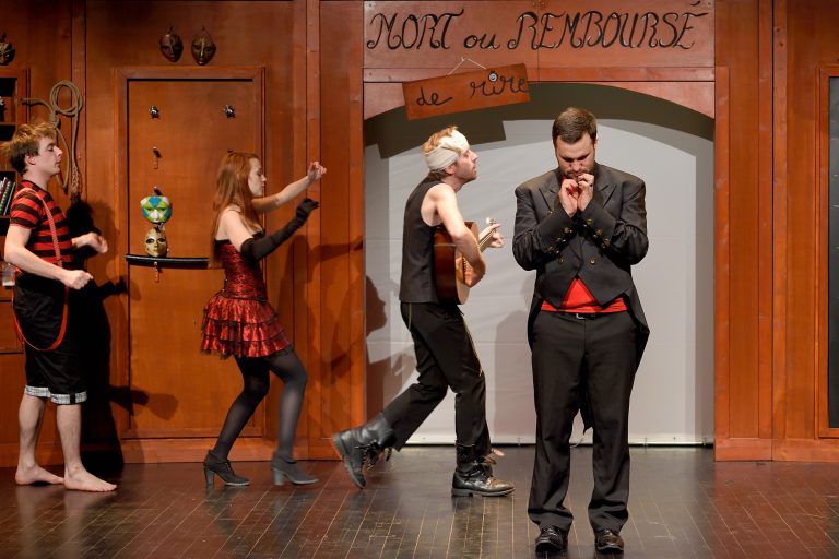 Image de la représentation "Le Magasin des Suicides" sur laquelle apparait un groupe de 3 acteurs entrain de jouer la pièce - une création de la compangie Nandi