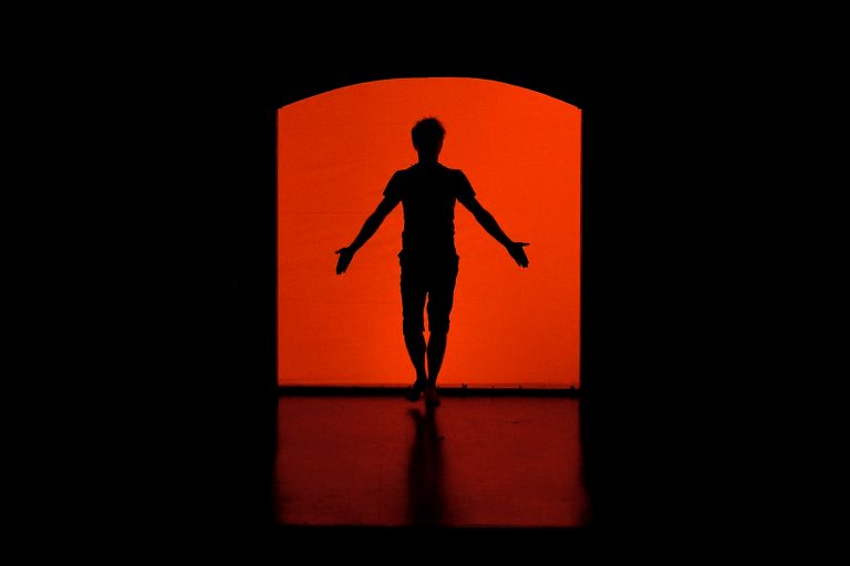 Image de la représentation "Le Magasin des Suicides" sur laquelle un acteur apparait sur un fond rouge et qui s'avance sur la scène - une création de la compangie Nandi