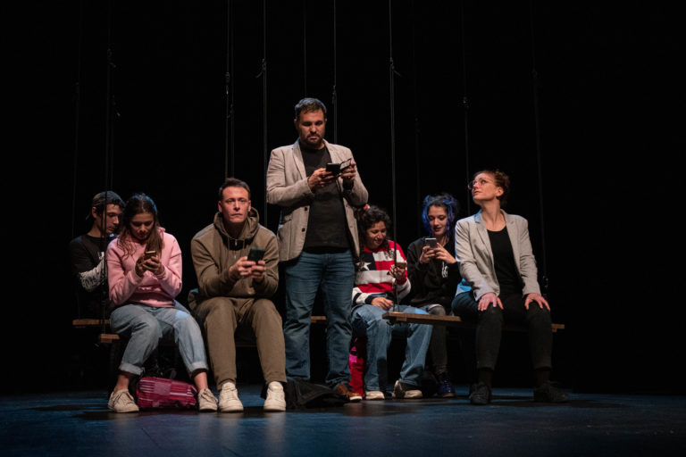 Image de la représentation "Michelle doit-on t'en vouloir d'avoir fait un selfie à Auschwitz ?" sur laquelle les acteurs sont tous assis derrière un autre tenant dans ses mains un micro - une création de la compagnie Nandi