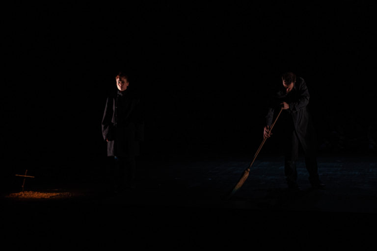 Image de la représentation "Michelle doit-on t'en vouloir d'avoir fait un selfie à Auschwitz ?" sur laquelle deux acteurs sont présents, l'un passant le balais, l'autre nous regardant droit dans les yeux - une création de la compagnie Nandi Lyon