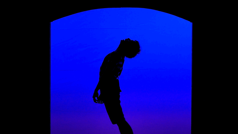 Scène centrée sur un acteur regardant le ciel, seul sur un fond bleu. Extrait de la représentation "Le Magasin des Suicides" de la compagnie de théâtre Cie Nandi Lyon